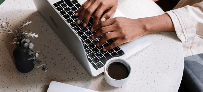 Hands typing on a laptop with a cup of coffee beside laptop